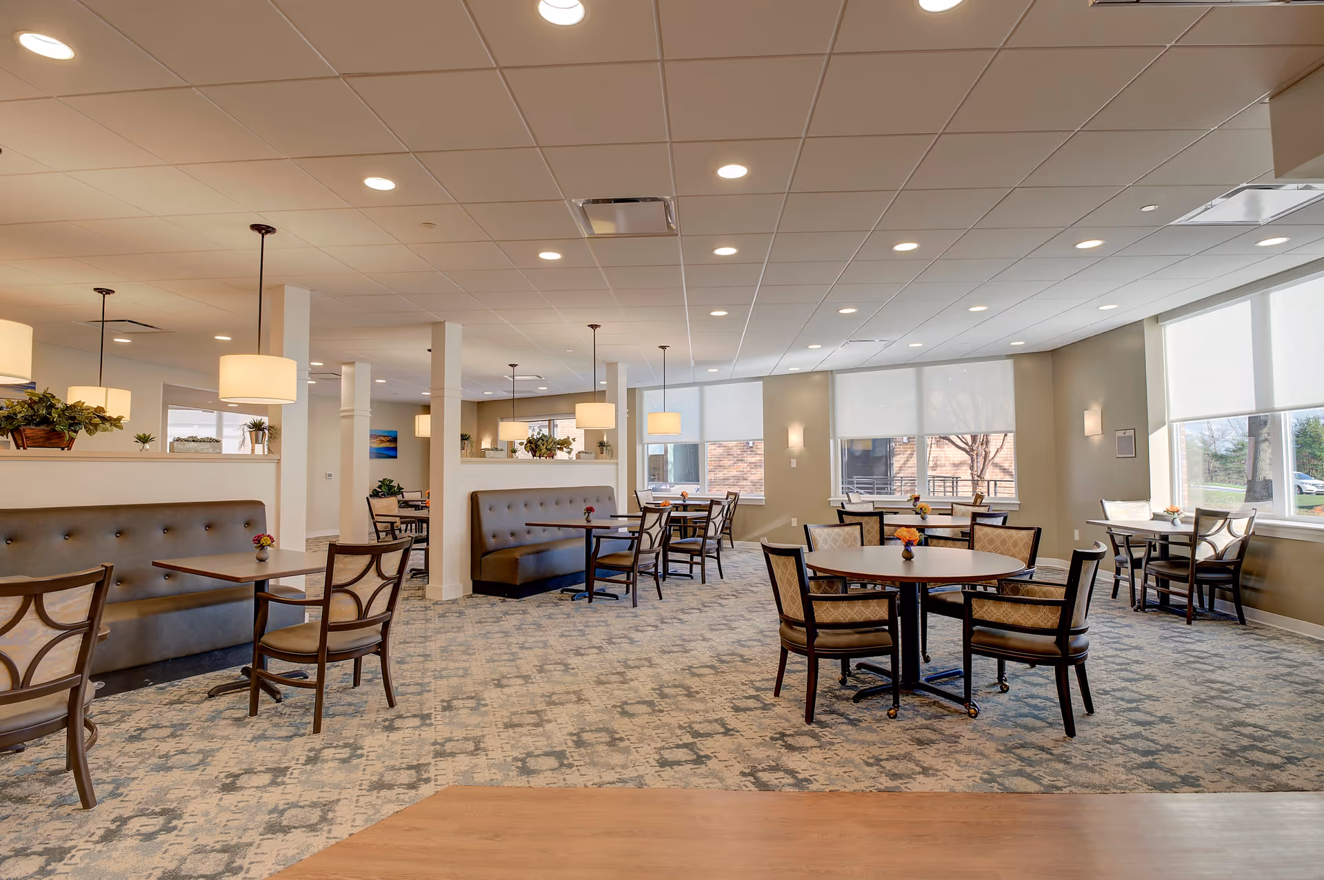 A bright and spacious dining room with multiple tables and chairs arranged neatly. The room features large windows with white roller shades allowing natural light to fill the space. There are cushioned bench seats along the walls and pendant lights hanging from the ceiling. Small flower arrangements are placed on each table, and the carpet has a patterned design.