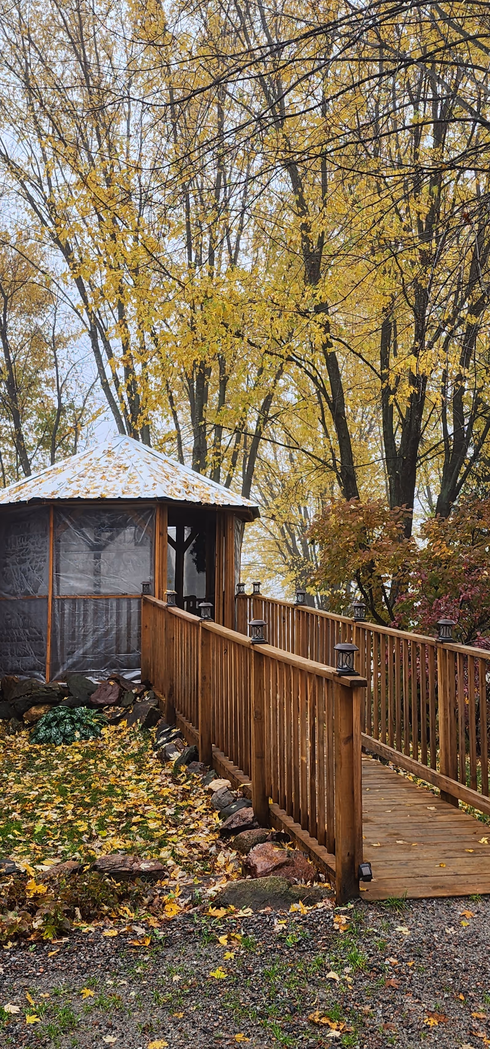 A wooden gazebo with a sloped roof covered in fallen leaves, surrounded by trees with yellow autumn foliage. A wooden ramp with railings and small lanterns on posts leads up to the gazebo. The ground is covered with fallen leaves and rocks line the edge of the ramp.