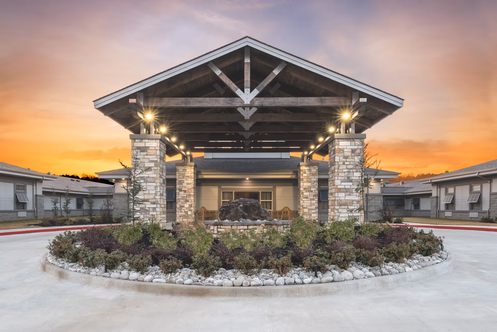 Entrance of Belvedere Nursing and Rehab facility at sunset, featuring a covered driveway with wooden beams and stone pillars, surrounded by landscaped plants and shrubs.