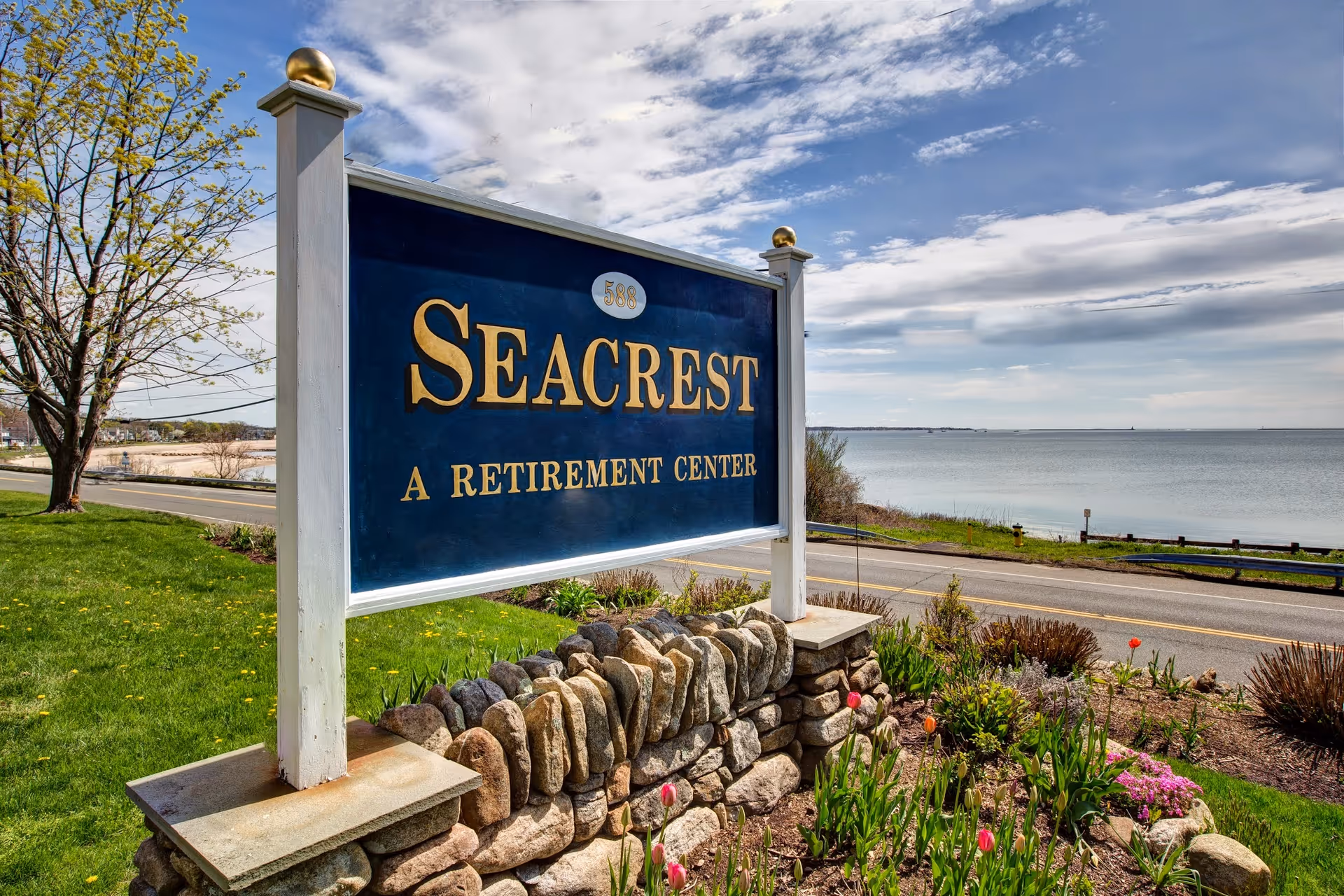 A blue sign reading "Seacrest A Retirement Center" mounted on a stone base beside a coastal road with the ocean in the background.