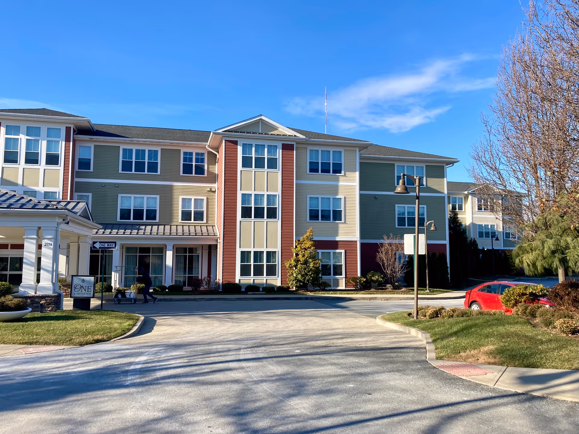 Front exterior of a three-story senior living building with a covered entrance, landscaped driveway, and a parked car.