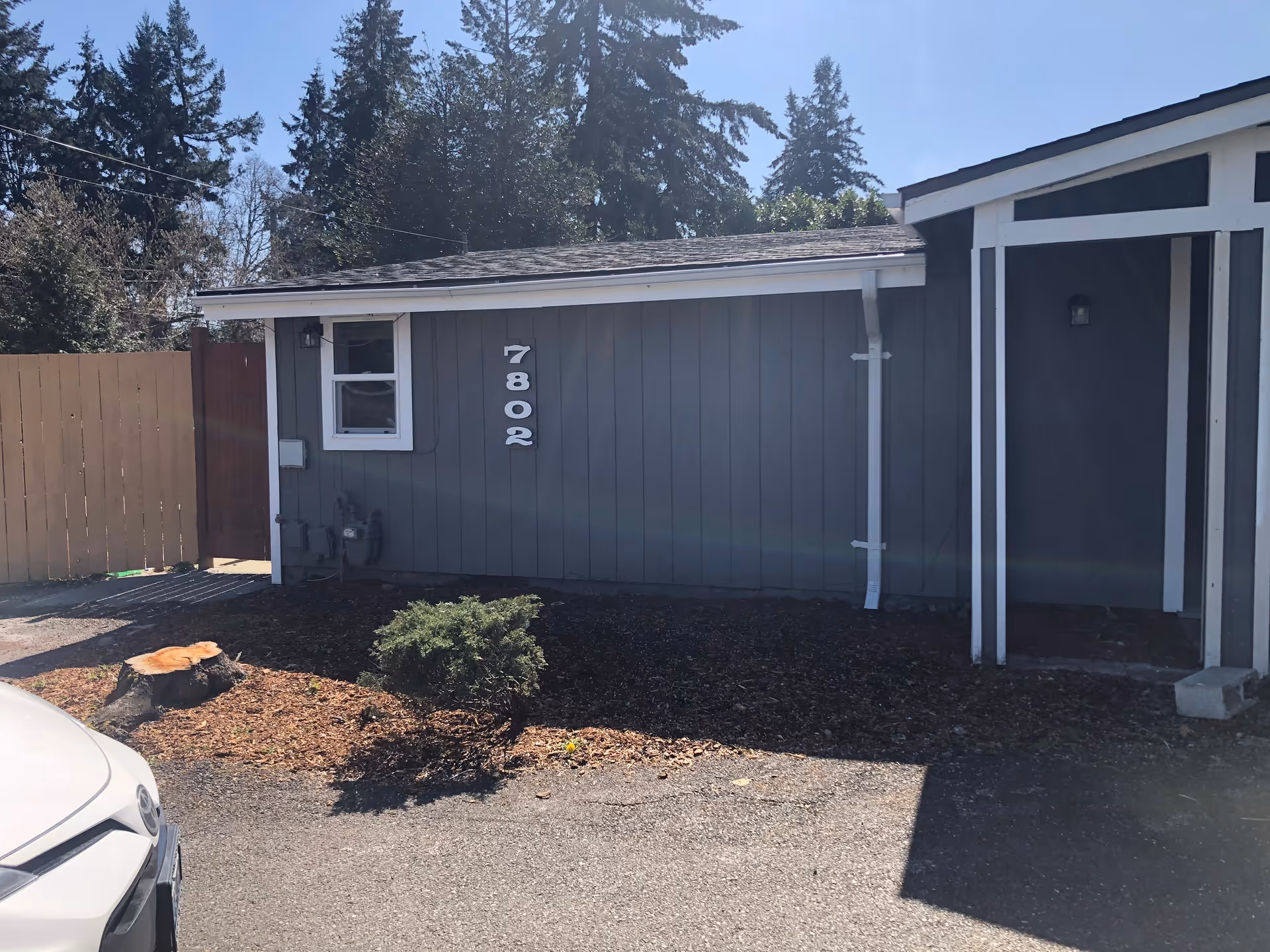 Exterior view of a gray building with white trim, showing the house number 7802 mounted on the wall. There is a small window, a wooden fence, a tree stump, and a small bush in front of the building. Part of a white car is visible in the lower left corner. Tall trees are in the background under a clear sky.