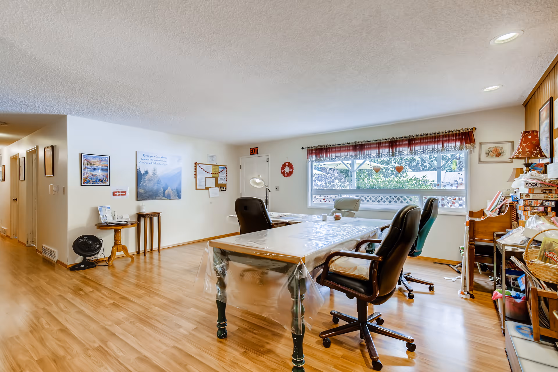Bright communal activity/dining room with long tables covered in plastic, rolling chairs, a large window and wood-look flooring.
