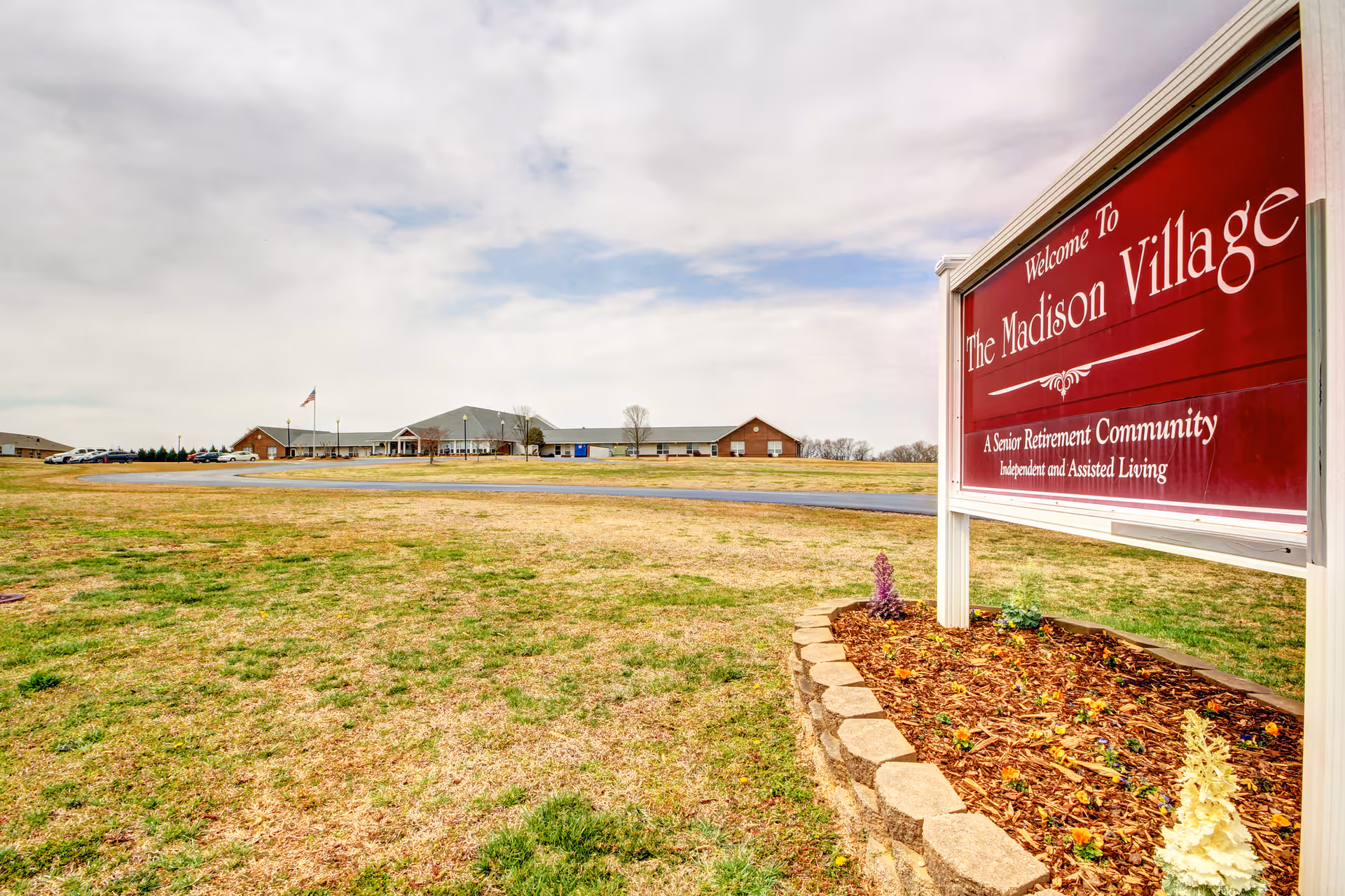 A red "Welcome to The Madison Village" sign in the foreground with the single-story retirement community building, lawn, and flagpole in the background.