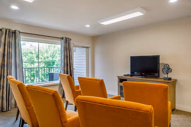 A cozy living room with four orange upholstered chairs arranged facing a wooden TV stand with a flat-screen television and decorative item. There is a large window with patterned curtains letting in natural light and a door leading to a balcony with outdoor seating visible.