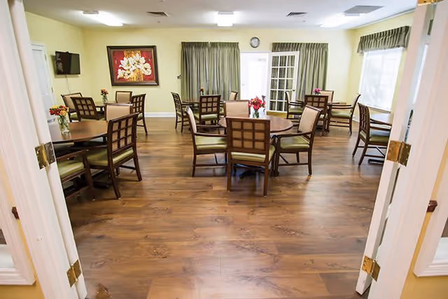 A dining room with multiple round wooden tables and cushioned chairs arranged neatly on a wooden floor. The room has light yellow walls, a wall-mounted TV, floral paintings, and windows with green curtains allowing natural light to enter.