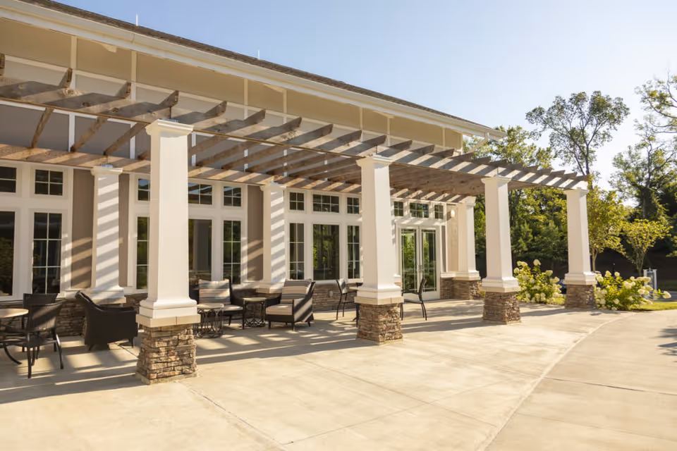 Outdoor patio area at The Heritage at Brentwood featuring a pergola with wooden beams supported by white columns with stone bases. The patio has several seating arrangements with chairs and small tables, surrounded by greenery and trees under a clear sky.