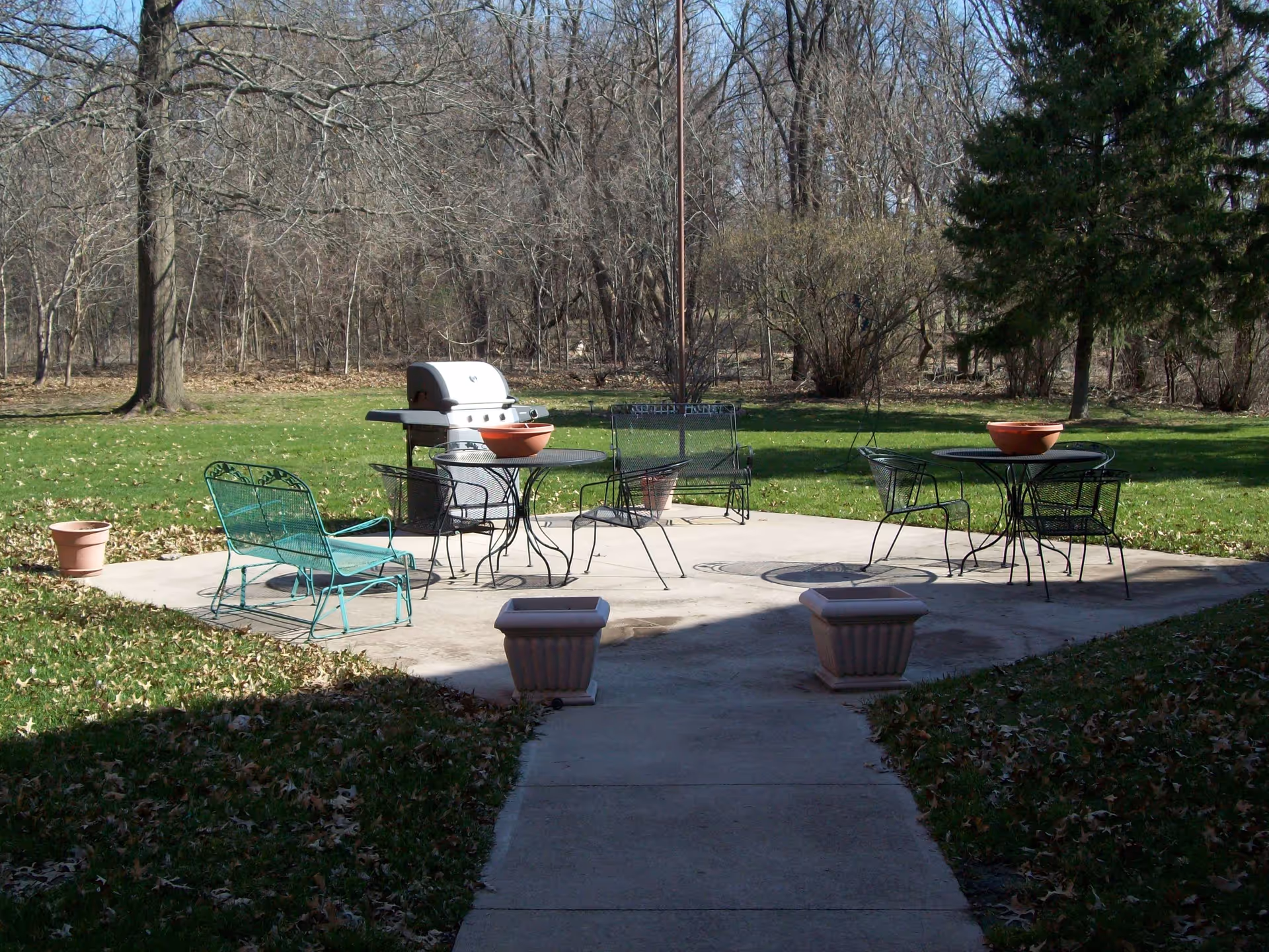Outdoor patio area with metal tables and chairs, a barbecue grill, and several empty flower pots on a concrete surface surrounded by grass and trees with bare branches.