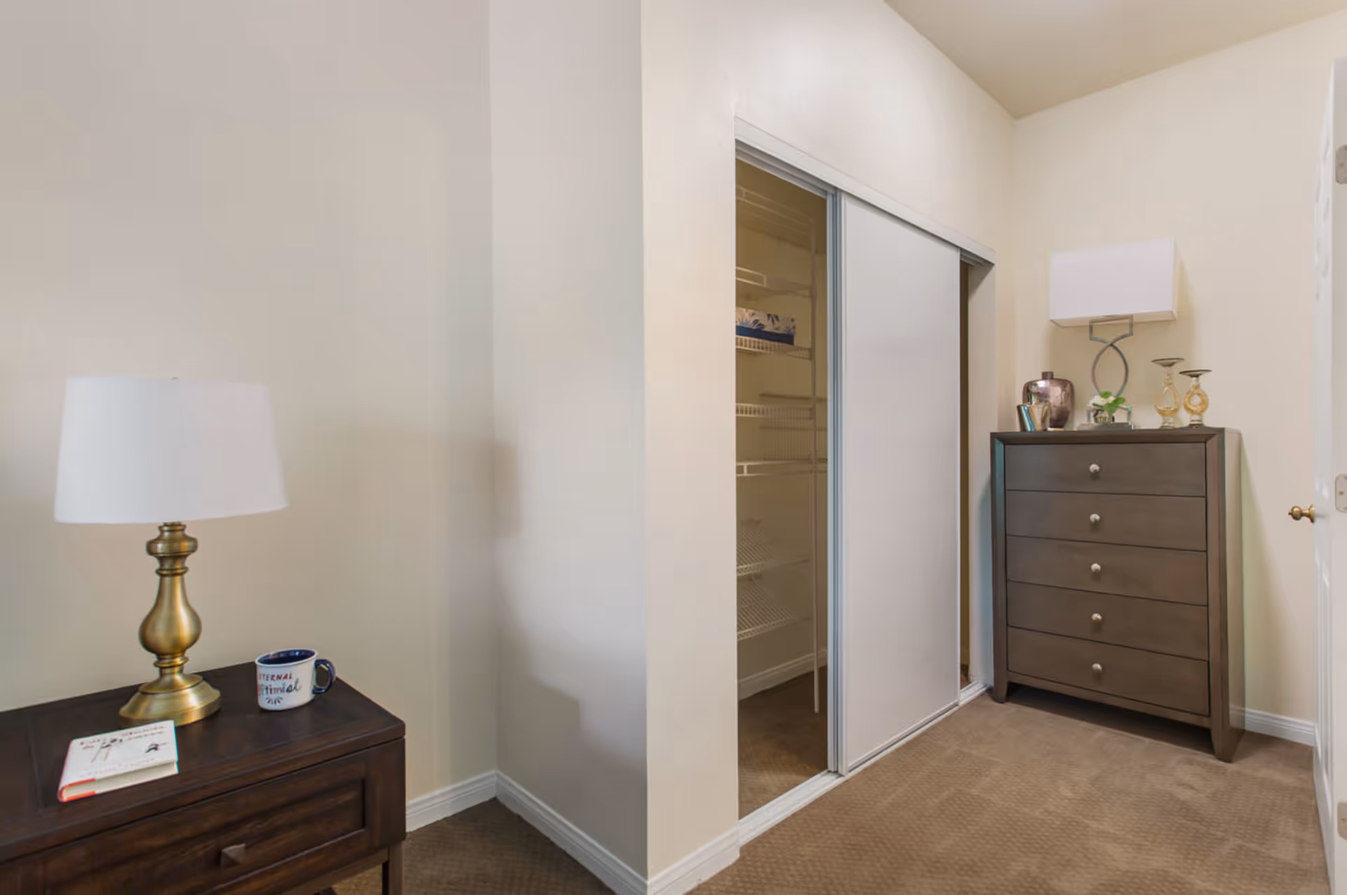 A small bedroom corner featuring a dark wooden nightstand with a brass lamp, a book, and a coffee mug on top. Adjacent to it is a closet with sliding doors, one door open revealing wire shelving inside. On the right side, there is a dark wooden chest of drawers with a modern lamp and decorative items on top. The walls are light-colored and the floor is carpeted.