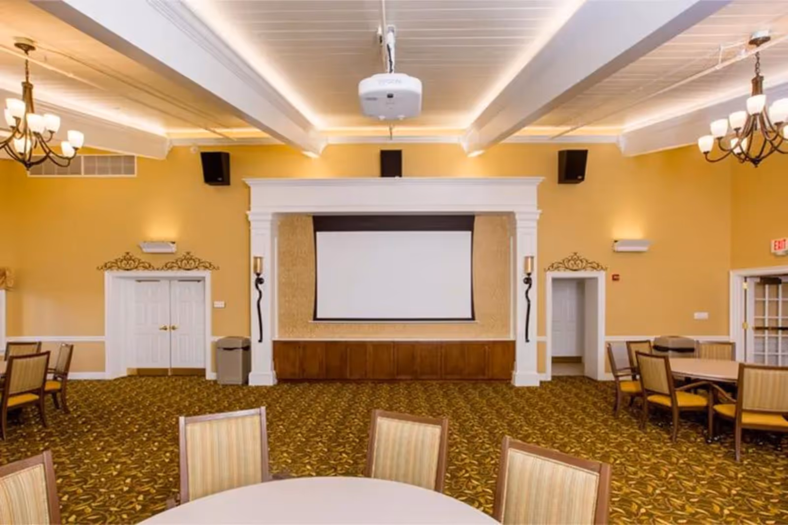 A spacious room with yellow walls and patterned carpet featuring multiple tables and chairs arranged for seating. The room has a large white projection screen mounted on a wall with decorative molding, two chandeliers hanging from the ceiling, and a ceiling-mounted projector. There are double doors and an exit door visible on the walls.