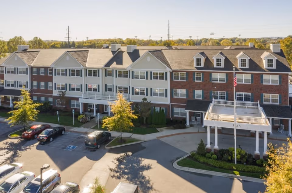 Exterior view of a three-story senior living facility building with a covered entrance, multiple windows, and a parking lot with several cars. Trees and greenery surround the building under a clear sky.