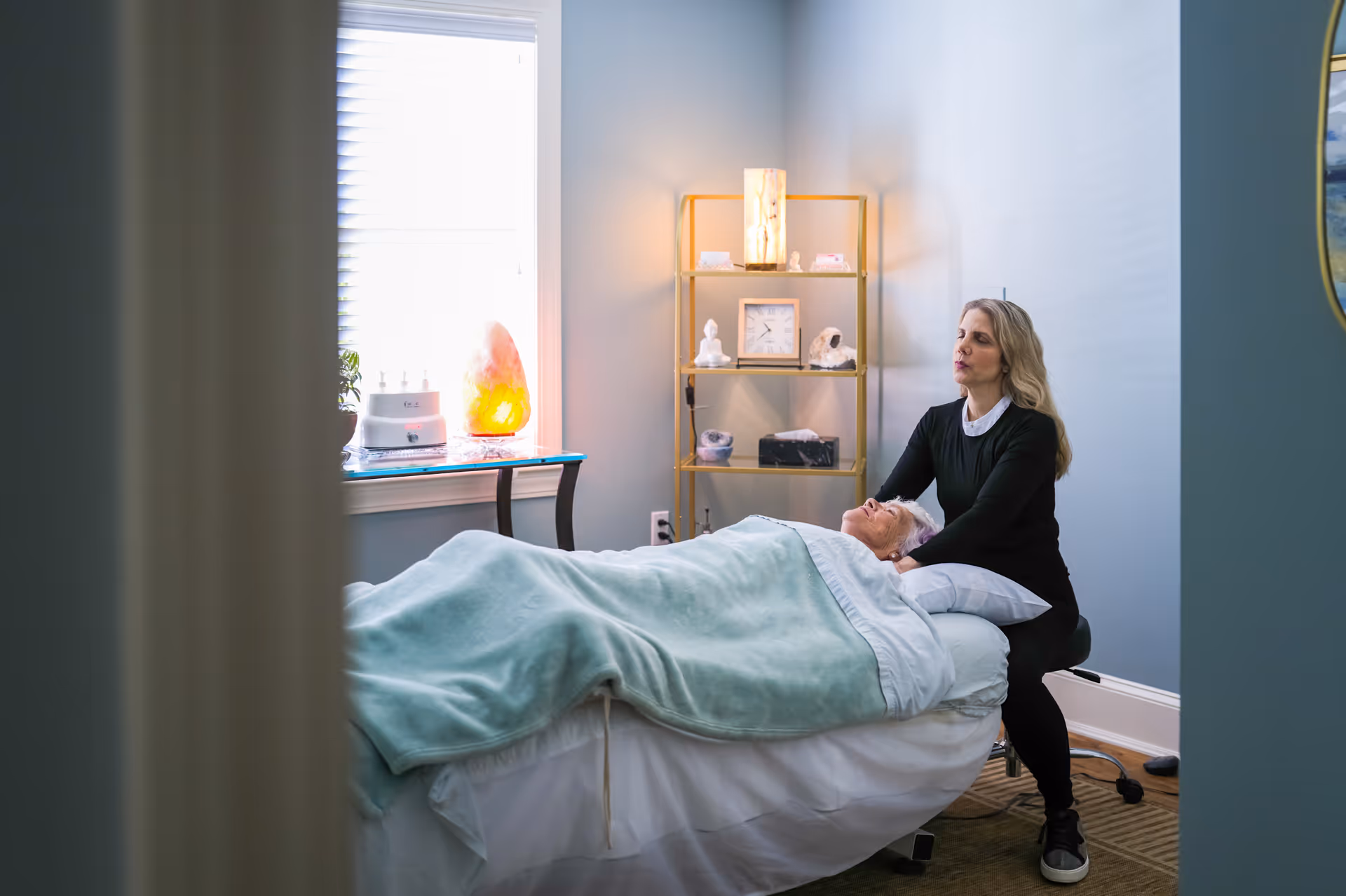 A woman is providing a therapeutic or healing treatment to an elderly person lying on a bed covered with a light green blanket in a softly lit room. The room has a window with blinds, a small table with a glowing salt lamp, and a gold-colored shelving unit with decorative items and a clock.