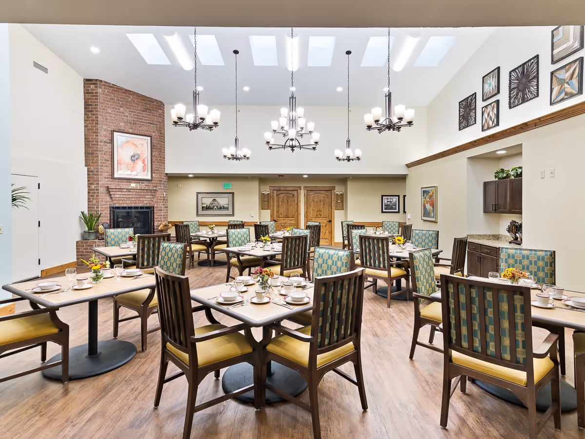 A bright dining room with multiple tables set for meals, each table surrounded by wooden chairs with patterned cushions. The room features a high ceiling with skylights and several chandeliers. There is a brick fireplace on the left wall with a framed artwork above it, and various framed pictures and decorative items on the walls. The floor is wooden, and the overall atmosphere is warm and inviting.