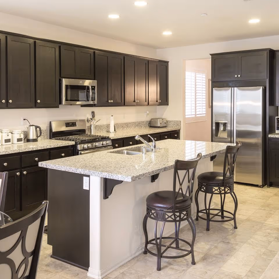 Modern kitchen with a large granite island, bar stools, dark cabinets, and stainless steel appliances.
