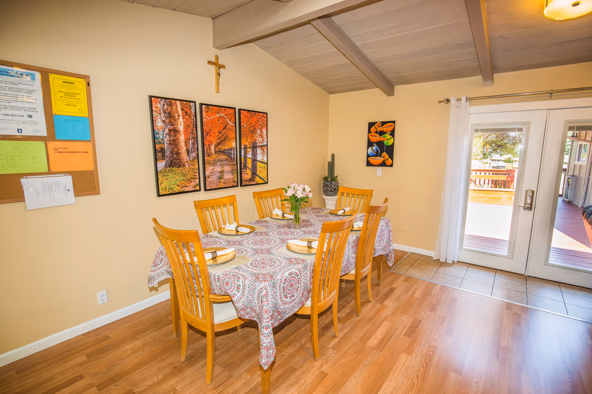 A dining room with a wooden table covered by a patterned tablecloth, set with six wooden chairs. The table is set with plates and napkins, and a vase with flowers is in the center. On the wall, there is a three-panel artwork depicting a tree-lined path with autumn foliage, a crucifix above it, and a smaller colorful painting. A bulletin board with various notices is on the left wall. Double glass doors with white curtains lead to an outdoor deck area. The floor is a combination of wood and tile.