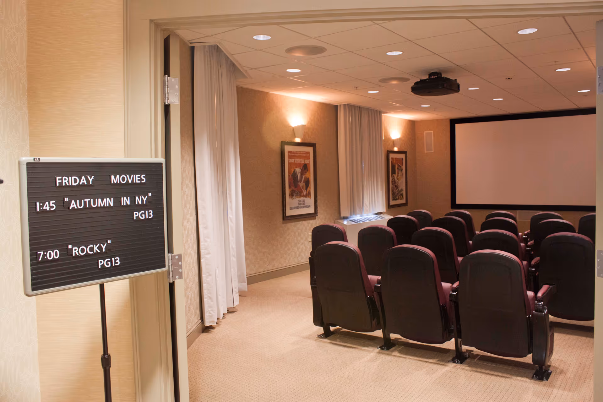 A small movie theater room with rows of maroon cushioned seats facing a large blank projection screen. On the left side, a sign displays Friday movie showtimes: 1:45 PM 'Autumn in NY' PG13 and 7:00 PM 'Rocky' PG13. The room has beige walls, carpeted floor, framed posters on the walls, and ceiling lights.