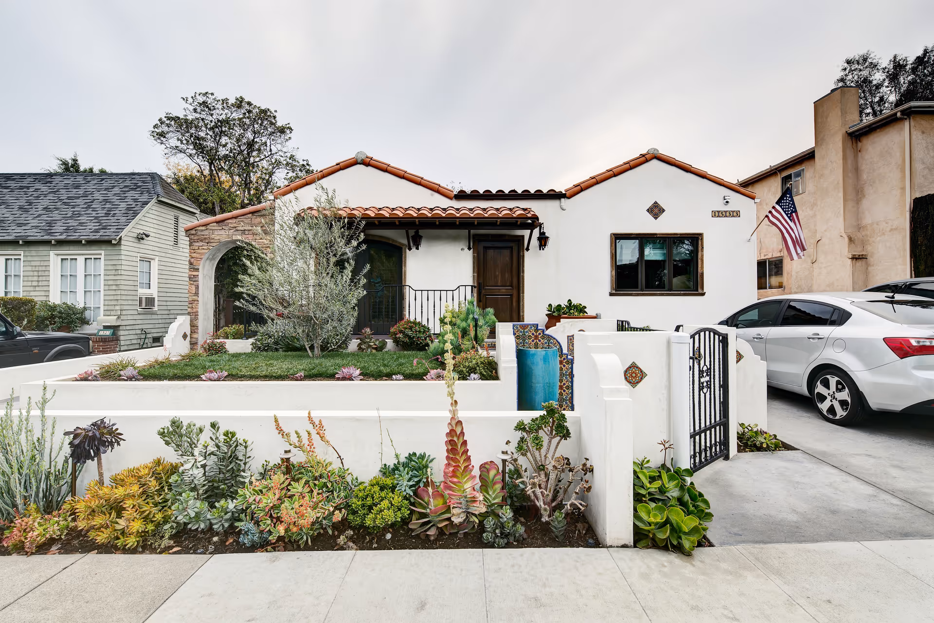 Front exterior view of a single-story house with white stucco walls and a red tile roof. The house has a small front yard with various succulents and plants, a gated driveway with a silver car parked, and an American flag mounted on the right side. The entrance features a wooden door and a small porch area with decorative tiles on the low walls.