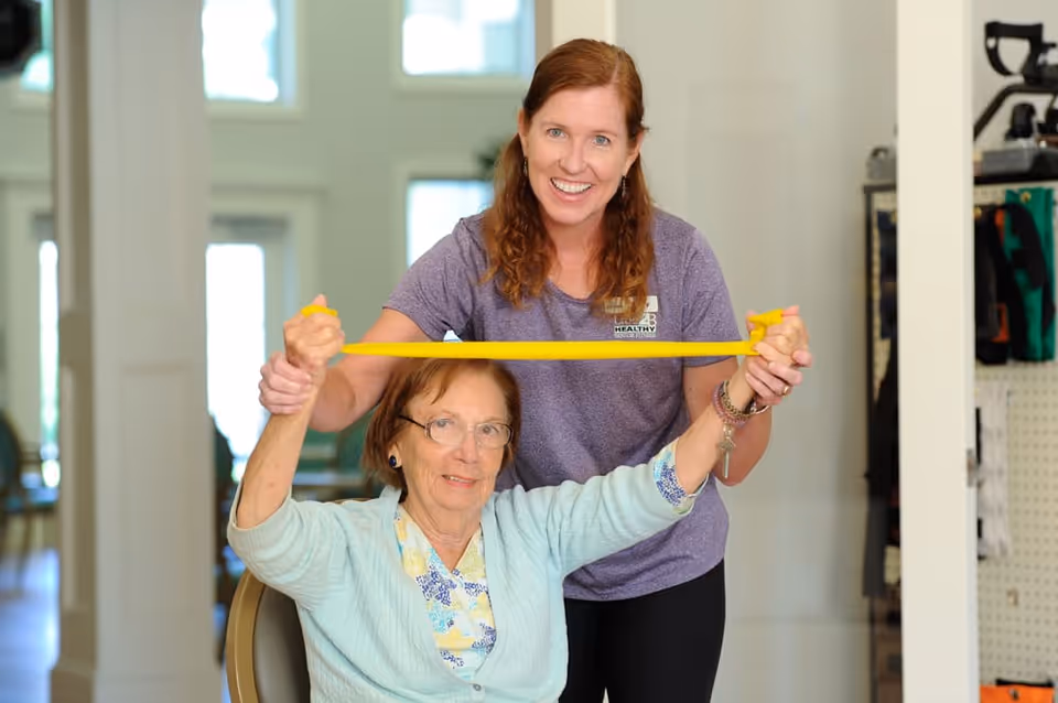 A senior woman sitting on a chair is assisted by a younger woman standing behind her as they stretch a yellow resistance band together in a bright room with large windows.