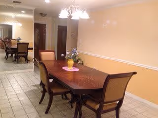 A dining area with a wooden dining table and six chairs. The table has a yellow vase with flowers on a pink doily. The room has tiled flooring and a two-tone wall painted in beige and white with a chair rail. In the background, there is another dining area with additional chairs and a chandelier overhead.
