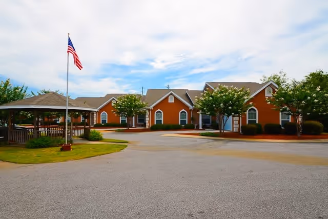 Exterior view of a single-story senior living facility building with a brownish-orange facade, white trim, and multiple arched windows. In front of the building is a paved driveway and a small grassy area with a gazebo and an American flag on a flagpole. Trees and shrubs are planted around the building and gazebo under a partly cloudy sky.