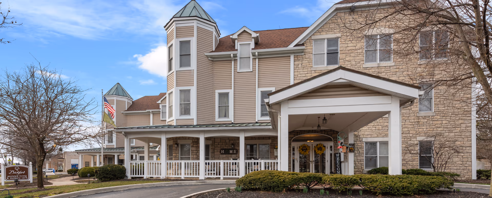 Exterior view of Bickford of Bexley, an assisted living and memory care facility, showing a large building with beige siding and stone accents, a covered entrance, and a porch with white railings. There are leafless trees and bushes around the driveway, and an American flag is visible near the building.