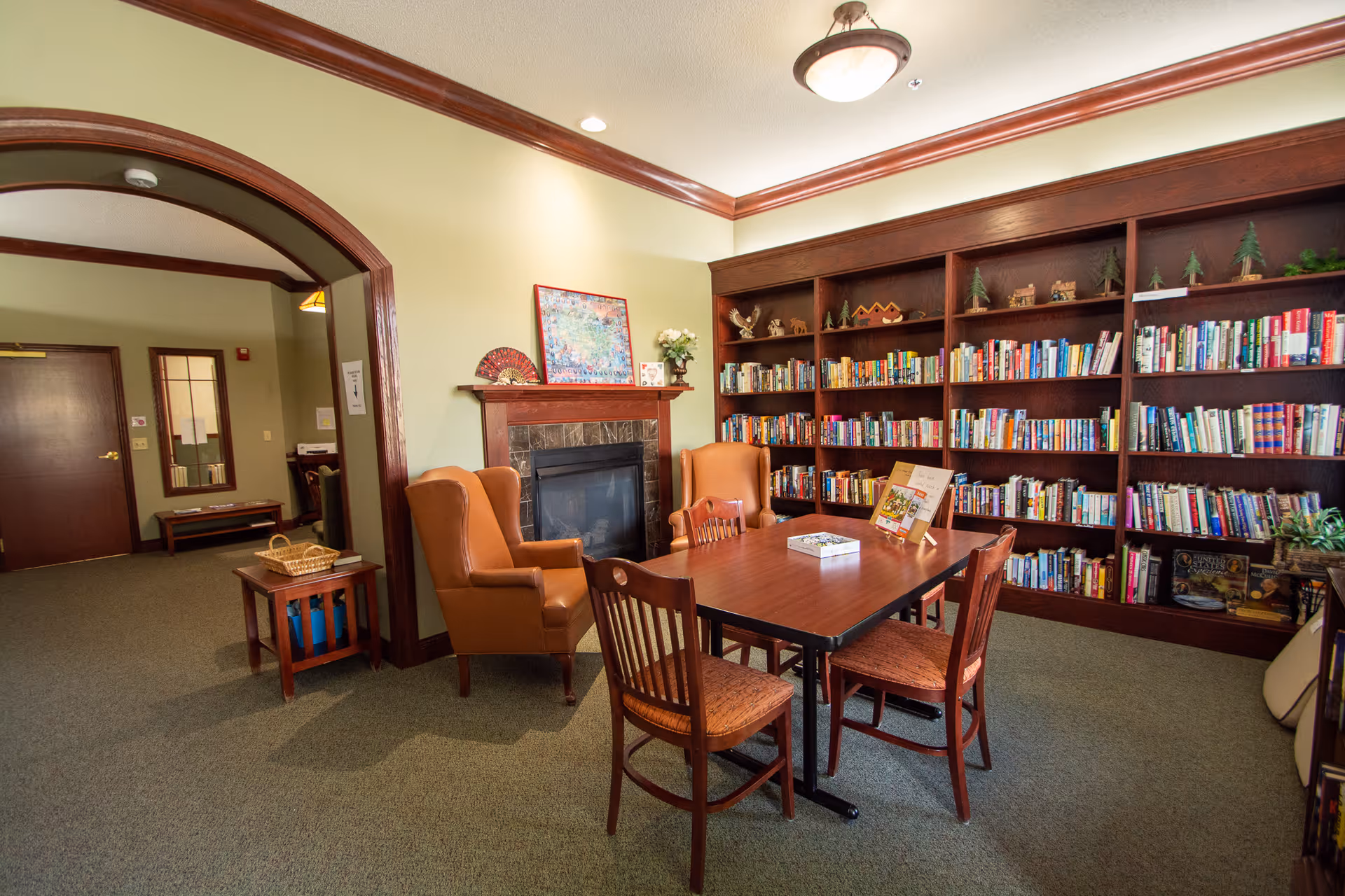 Cozy library-style common room with bookshelves, a table and chairs, two armchairs, and a fireplace.