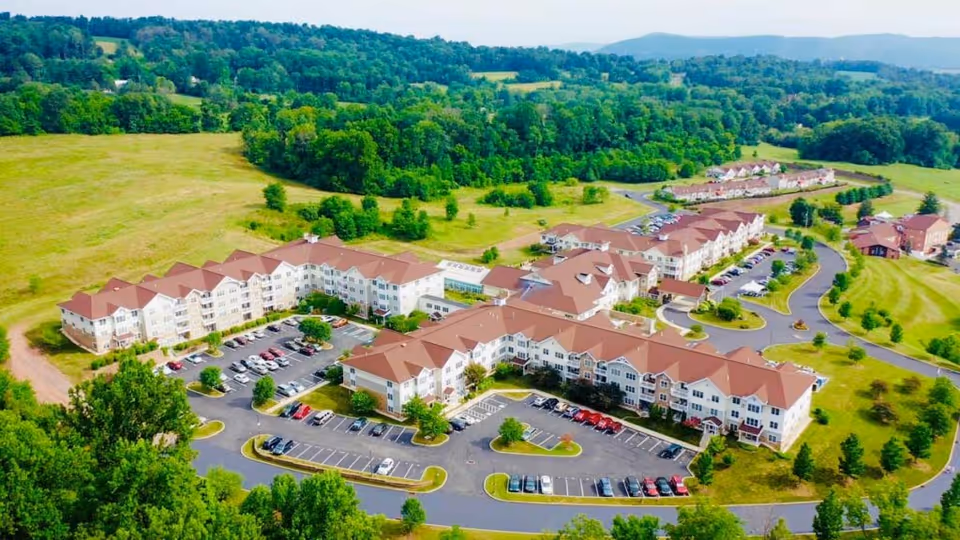 Aerial view of The Heritage Of Green Hills senior living facility showing multiple connected buildings with red roofs surrounded by parking lots, green lawns, trees, and rolling hills in the background under a clear sky.