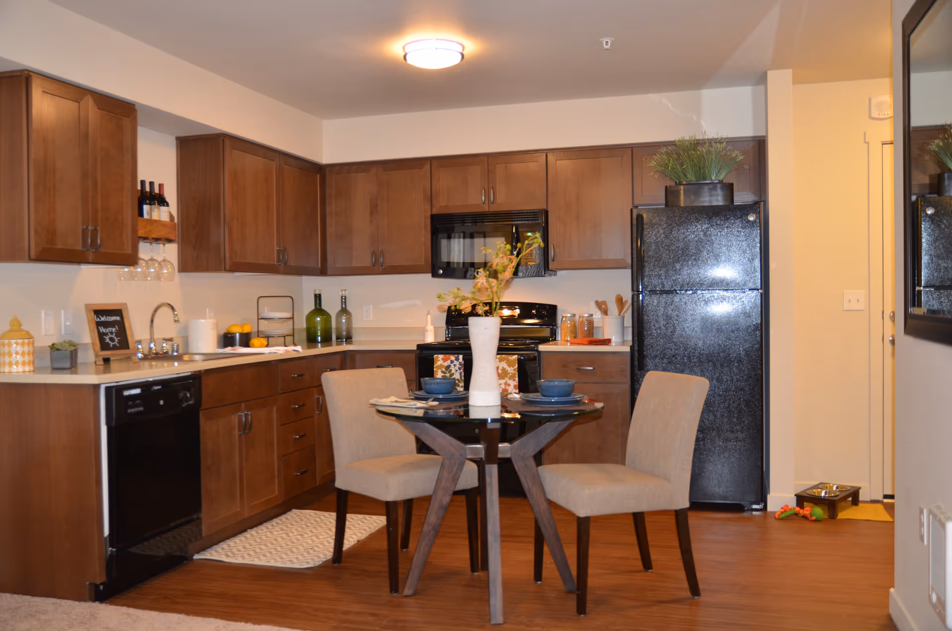 A modern kitchen and dining area with wooden cabinets, black appliances including a refrigerator, stove, microwave, and dishwasher. A small round dining table with two beige upholstered chairs is set with blue bowls and plates, and a white vase with flowers is in the center. The kitchen counter has decorative items including bottles, a small chalkboard sign, and a plant. The floor is wooden, and a mirror is visible on the right wall.