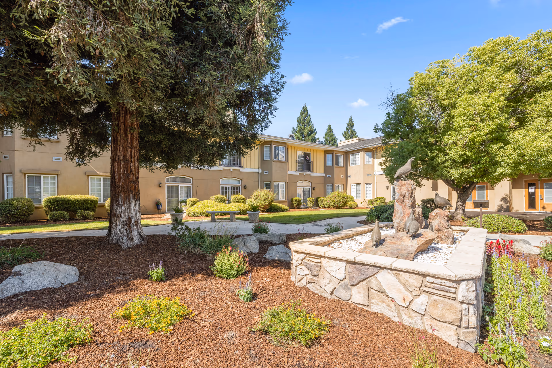 Outdoor courtyard area of a senior living facility with a large tree, landscaped garden beds, a stone planter with bird sculptures, and a two-story building in the background under a blue sky with a few clouds.