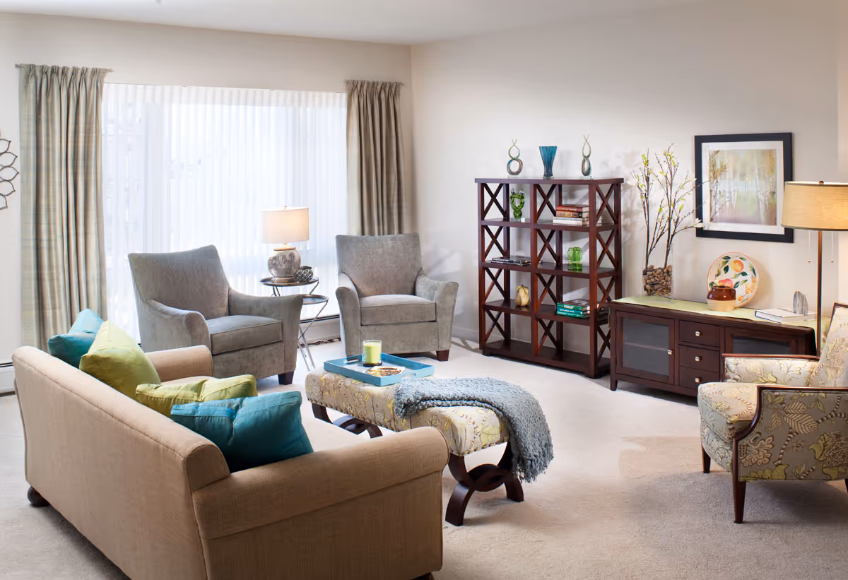 A bright and cozy living room with a beige sofa adorned with green and blue pillows, two gray armchairs, and a patterned armchair. There is a cushioned ottoman with a blue throw blanket and a tray on top. The room features a wooden bookshelf with decorative items and books, a wooden sideboard with a lamp, framed artwork, and a vase with branches. Large windows with sheer curtains allow natural light to fill the space.