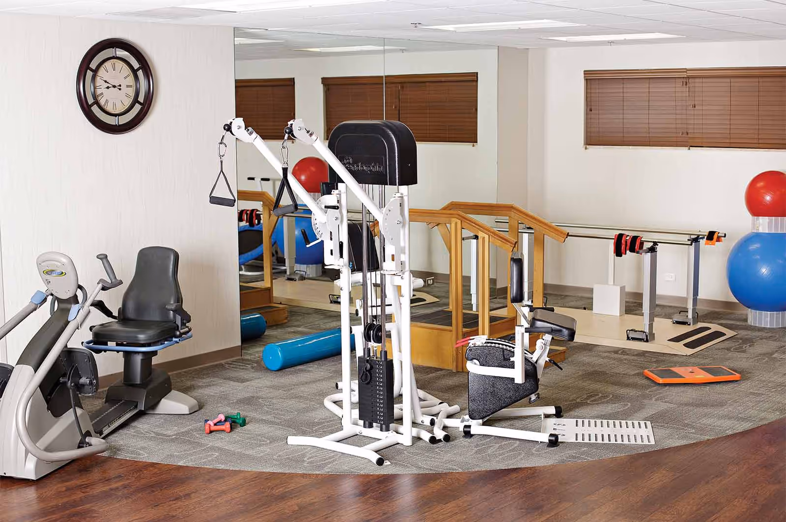 An indoor fitness/physical therapy room with exercise machines, balance balls, handrails, and small weights.
