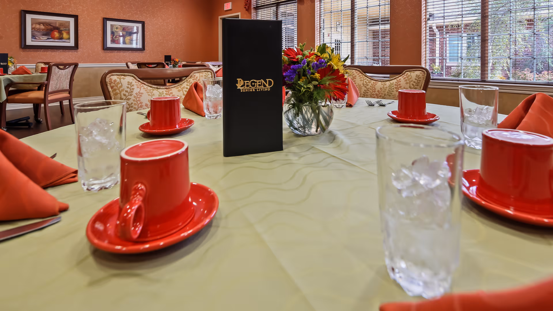 A dining table set with red cups and saucers, glasses of ice water, and red folded napkins in a senior living facility dining room. A black menu with the text 'Legend Senior Living' is placed in the center of the table along with a vase of colorful flowers. The room has large windows with blinds and upholstered chairs around the tables.