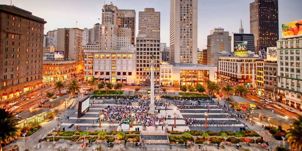 Aerial view of a large urban plaza with a tall central monument surrounded by people sitting and walking. The plaza is bordered by tall buildings with illuminated windows and palm trees lining the area. The scene is captured during dusk with city lights beginning to glow.
