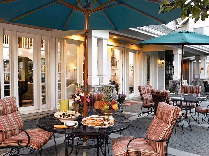Outdoor patio area with round metal tables and striped cushioned chairs under large green umbrellas. One table is set with plates of food, a pitcher, and a fruit bowl. The patio is adjacent to a building with large windows and warm interior lighting.