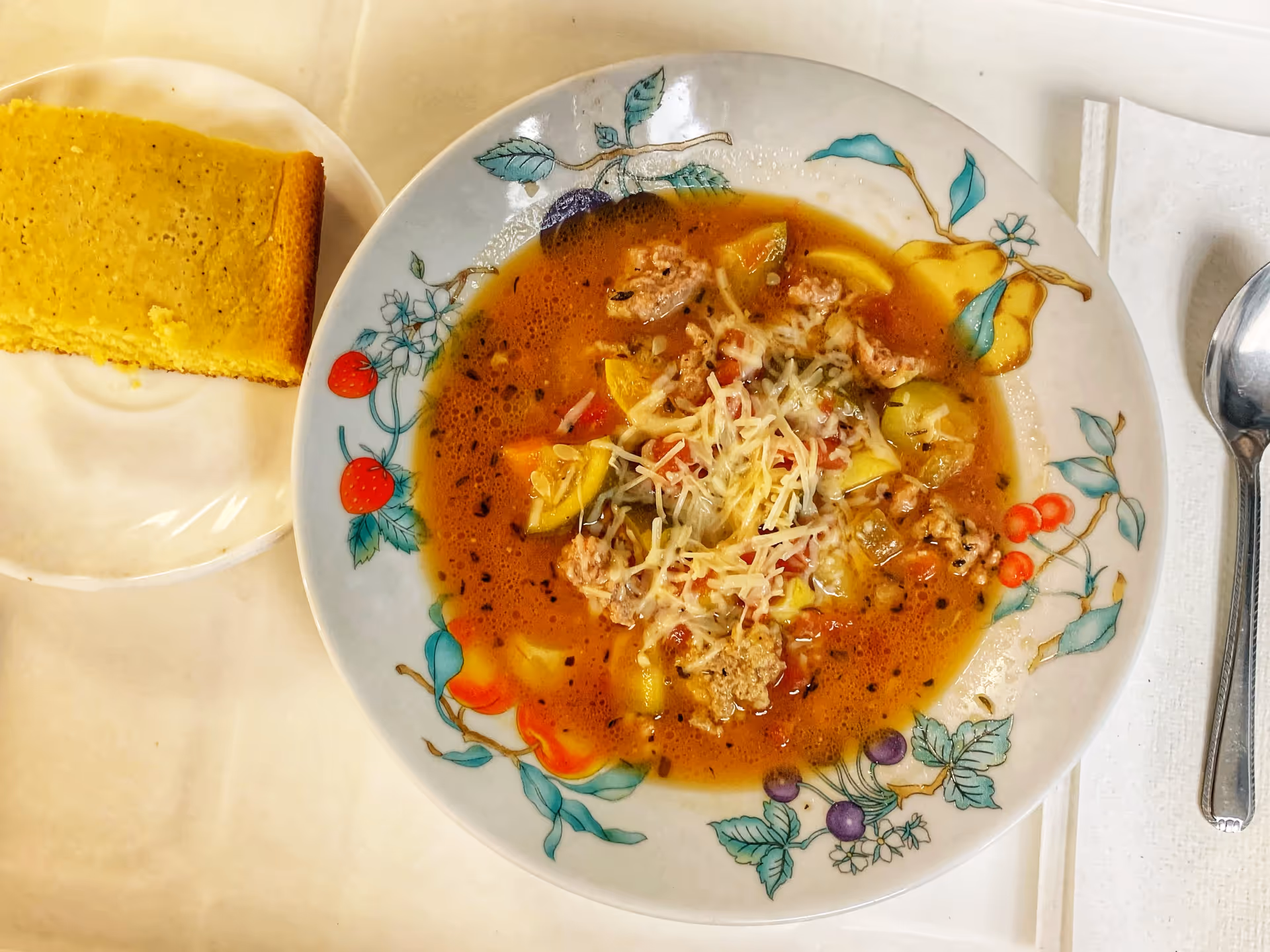 A bowl of vegetable soup with ground meat and shredded cheese on top, served on a decorative plate with fruit designs. Next to the bowl is a piece of cornbread on a white plate, a spoon, and a folded white napkin on a light-colored table.
