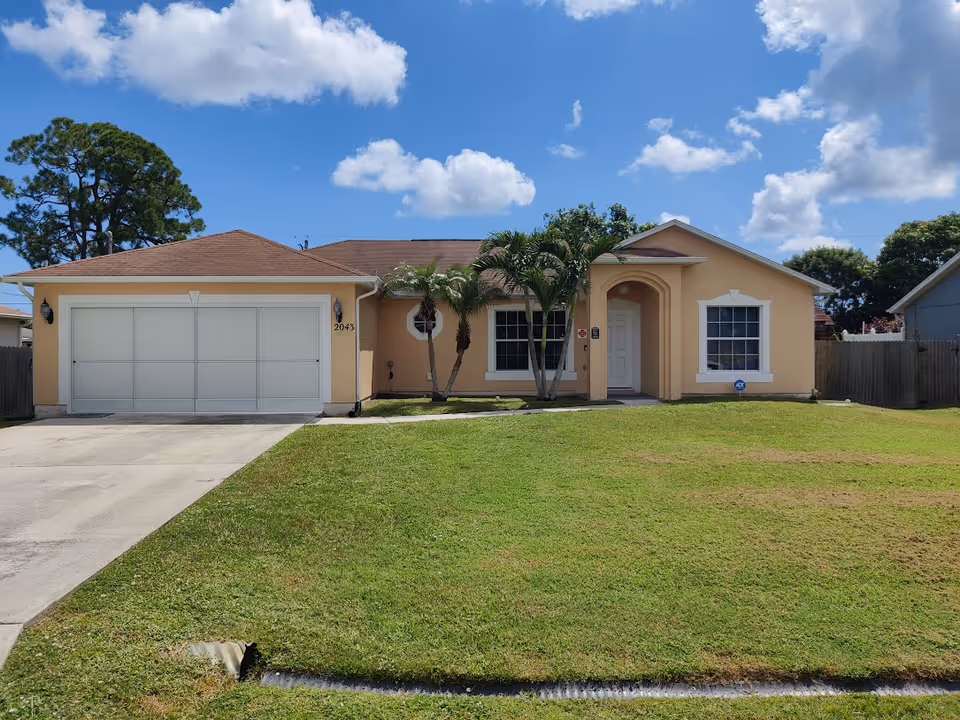 Front exterior view of a single-story peach-colored house with a brown roof, a two-car garage, a driveway, and a well-maintained lawn. There are two palm trees near the front entrance and a clear blue sky with some clouds above.