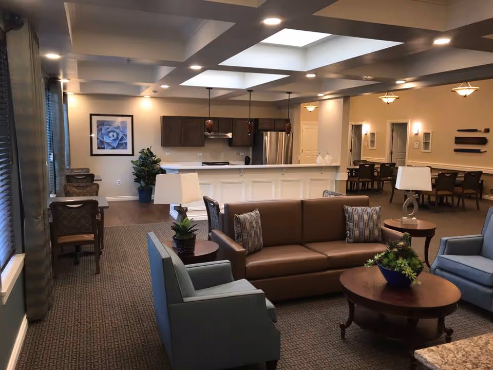 A spacious and well-lit interior common area featuring a brown leather sofa with patterned pillows, two blue armchairs, a round wooden coffee table with a plant centerpiece, and side tables with lamps. In the background, there is a kitchen area with a white island counter, dark wooden cabinets, stainless steel refrigerator, and pendant lights. Along the left wall, there are several desks and chairs near large windows with curtains. The room has a carpeted floor and recessed lighting in the ceiling.