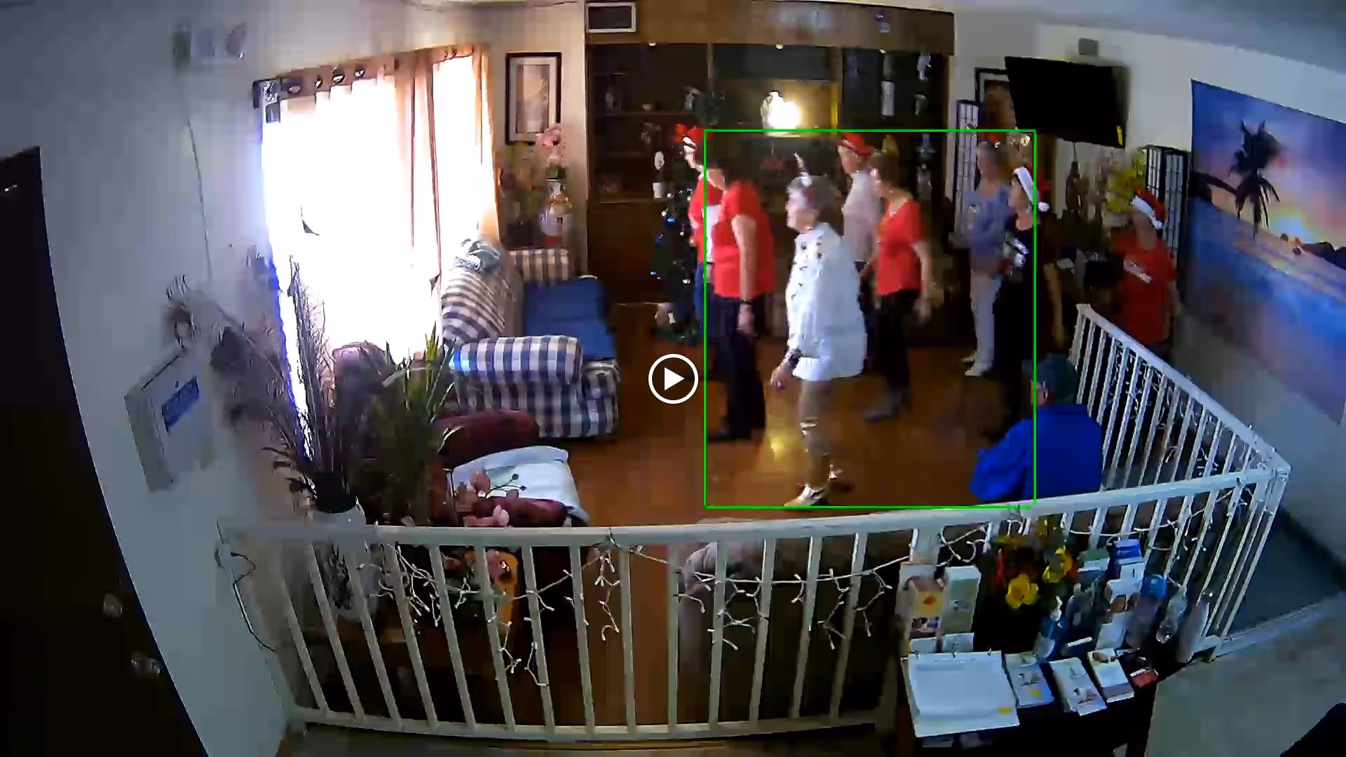 A cozy living room area in Foothills Adult Care Home with several elderly people standing and interacting. The room features a checkered armchair, a Christmas tree, a large wall-mounted TV, and a tropical beach painting on the wall. There is a white safety gate separating the seating area from a small table with various items and flowers.