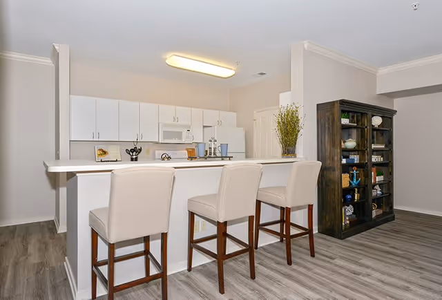 A modern kitchen area with white cabinets, a white countertop island with three beige cushioned bar stools, a microwave, and a refrigerator. To the right, there is a dark wooden bookshelf filled with decorative items. The floor has a light wood finish and the walls are painted light beige.