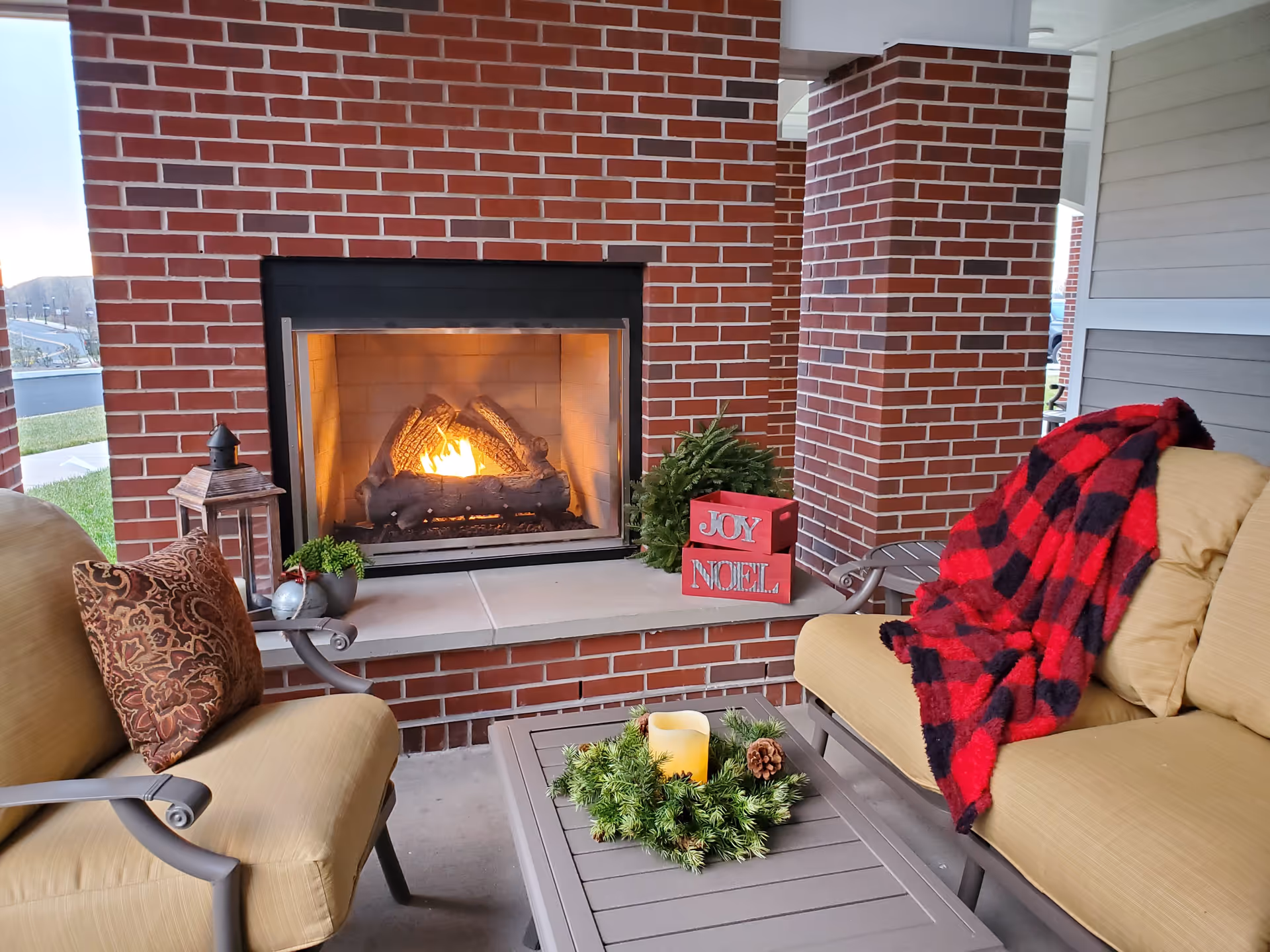 Outdoor seating area with cushioned chairs and a table in front of a brick fireplace with a fire burning. The table has a candle surrounded by a green wreath. A red and black checkered blanket is draped over one chair, and a decorative pillow is on another. There are holiday decorations including a small wreath and red blocks with the words JOY and NOEL on the fireplace ledge.