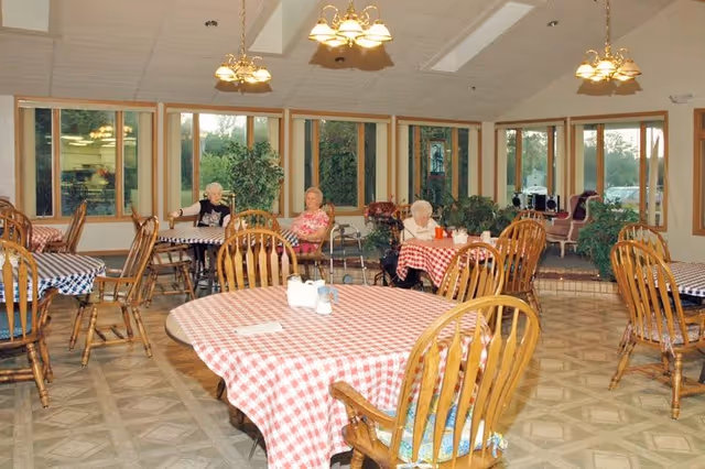 A dining room with several wooden tables covered with red and white checkered tablecloths. There are wooden chairs around the tables. Three elderly women are seated at different tables, and large windows line the walls, letting in natural light. The room has hanging light fixtures and some potted plants near the windows.