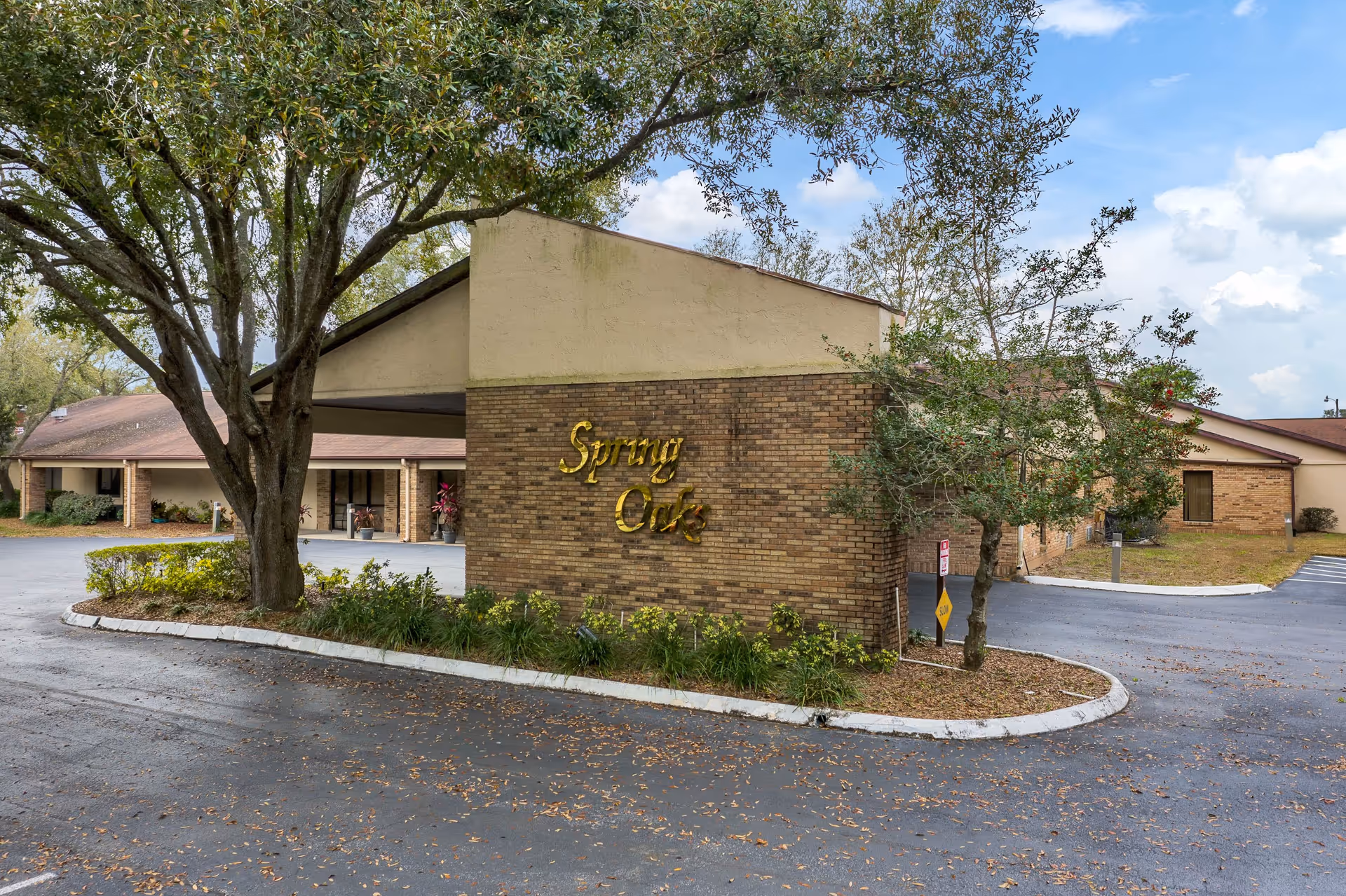 Exterior view of a senior living facility named Spring Oaks, showing a brick building with a covered entrance, surrounded by trees and a paved driveway.