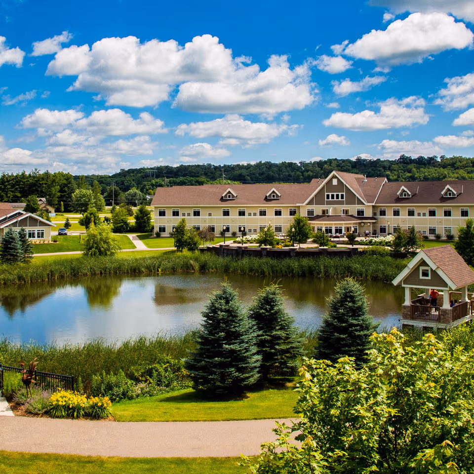 A large senior living facility building with beige siding and a brown roof, situated behind a pond surrounded by green grass, trees, and shrubs. There is a small gazebo on the right side near the pond with people inside. The sky is bright blue with scattered white clouds.