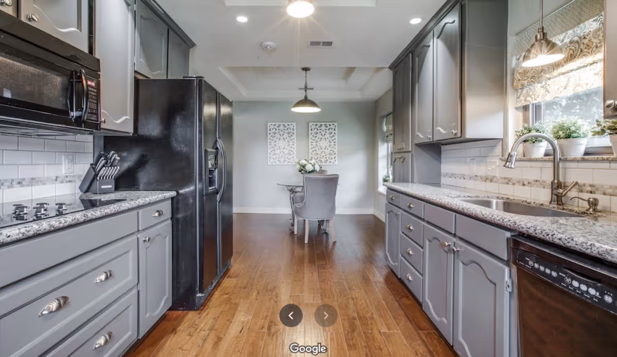 A modern kitchen with gray cabinets, granite countertops, a black refrigerator, microwave, stove, and dishwasher. The kitchen has wooden flooring and a dining area with a table and two chairs at the far end. There are two decorative wall hangings above the dining table and a window with a patterned Roman shade above the sink.