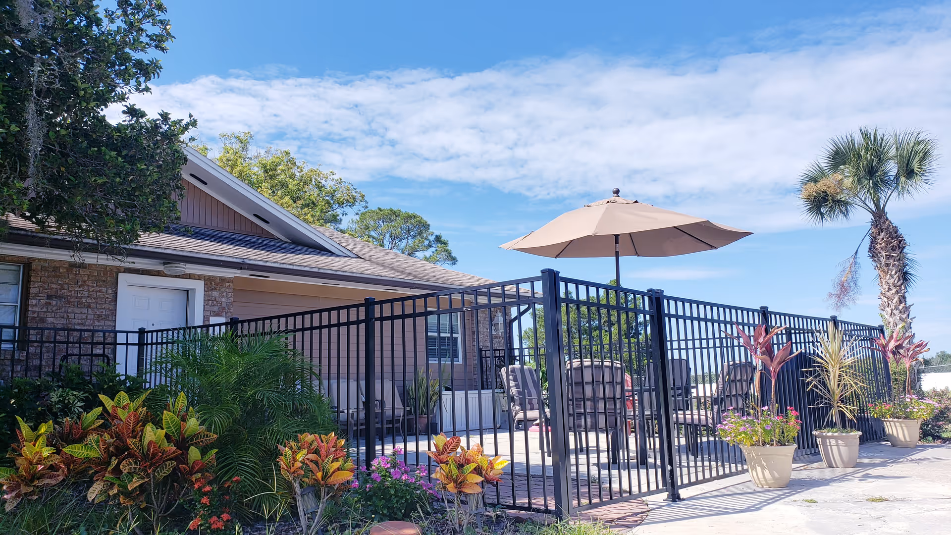 Outdoor patio area at Pinedale Gardens Assisted Living with a black metal fence surrounding a seating area that includes chairs and a large beige umbrella. The patio is decorated with potted plants and colorful flowers, with a palm tree and other greenery visible. The building exterior is brick and wood siding under a partly cloudy blue sky.
