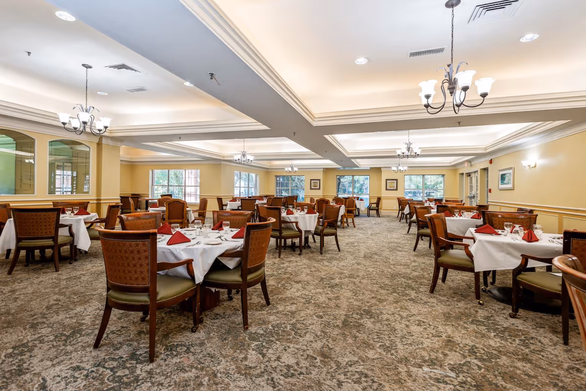 A spacious dining room with multiple round tables covered with white tablecloths and set with glassware and red folded napkins. The room has beige walls, large windows letting in natural light, carpeted floors, and elegant chandeliers hanging from a coffered ceiling.