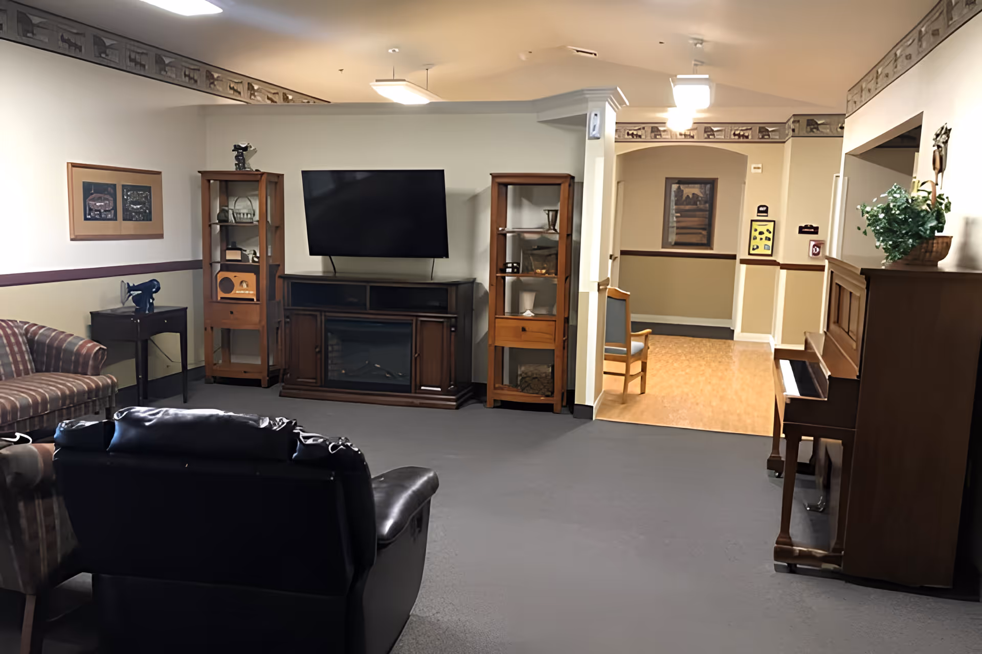 A cozy living room area in a senior living facility featuring a flat-screen TV mounted above a wooden cabinet with a fireplace insert. The room includes a striped couch, a black leather recliner, two wooden display cabinets with decorative items, a small side table with a vintage radio, and a wooden piano with a plant on top. The walls are decorated with framed pictures and a wallpaper border near the ceiling. A hallway with a chair and artwork is visible in the background.