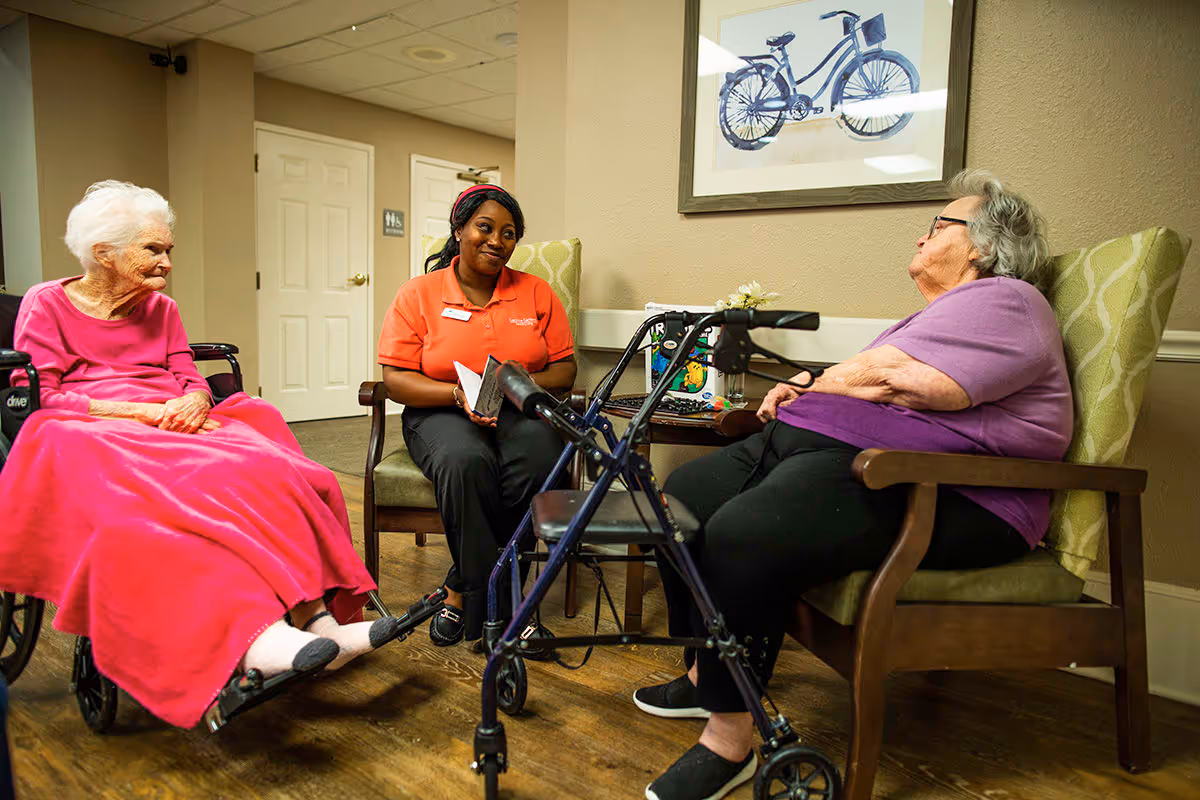 Two elderly women sitting in chairs in a common area of a senior living facility, one using a walker and the other in a wheelchair, with a staff member in an orange shirt sitting between them holding a notebook. A framed picture of a bicycle hangs on the wall behind them.