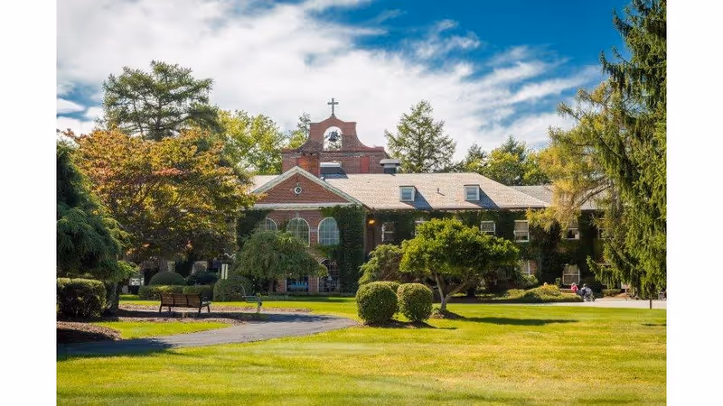 Front view of a brick nursing home building with a cross-topped cupola, surrounded by manicured lawns, trees, and a bench under a partly cloudy sky.