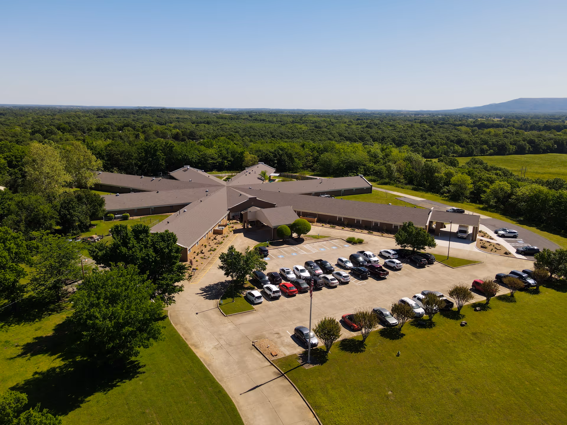 Aerial view of Oaks Healthcare Center showing a star-shaped single-story building with a parking lot, cars, and surrounding trees and fields.